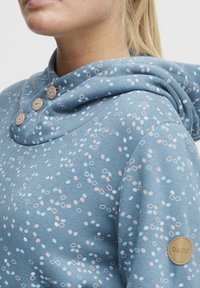 Sudadera azul con un diseño estampado de círculos rosas y blancos, que cuenta con una gran capucha, tres botones de madera y una etiqueta de Oxmo en la manga.