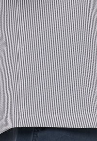 Pamučna srajca z oznako, ki prikazuje vertikalne sive in bele črte, rahlo ukrivljen rob ter vidna šivanja ob robovih.