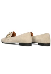 Beige suède loafers met een platte zwarte zool, voorzien van een decoratieve metalen gesp aan de voorkant en een glad leren interieur.