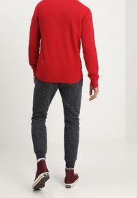 Pull en tricot rouge, pantalon de jogging gris foncé, baskets bordeaux. Coupe ajustée, poignets côtelés, tissu texturé et détails de couleur contrastée sur les chaussures.
