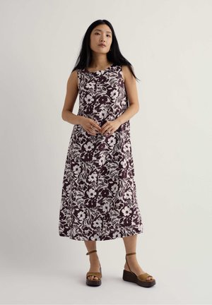 Žena s dlhými čiernymi vlasmi, ktorá má na sebe bezrukávové kvetinové šaty po členky a hnedé platformové sandále, stojí pred jednofarebným pozadím.