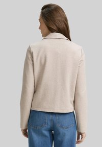 Beige, textuurjasje met een reverskraag, lange mouwen en een cropped lengte. Gecombineerd met high-waisted blauwe jeans met achterzakken.