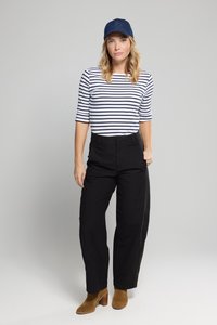 Kvinde iført mørkeblå kasket, hvid- og sortstribet skjorte, sorte brede bukser og brune suedestøvler, stående mod en grå baggrund.