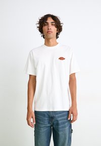 Hvid bomulds t-shirt med korte ærmer, rund hals og et lille farvet logo på venstre bryst. Båret sammen med blå denim jeans.