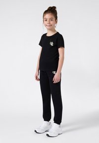 Κορίτσι που στέκεται με μαύρη μπλούζα και φόρμες, φορώντας λευκά αθλητικά παπούτσια, χαμογελώντας, μπροστά σε έναν ανοιχτό λευκό φόντο.