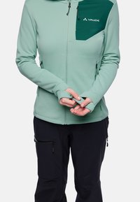 Giacca zip-up verde chiaro con una tasca sul petto di un verde scuro a contrasto, dotata di fori per il pollice, realizzata in un materiale morbido ed elasticizzato.