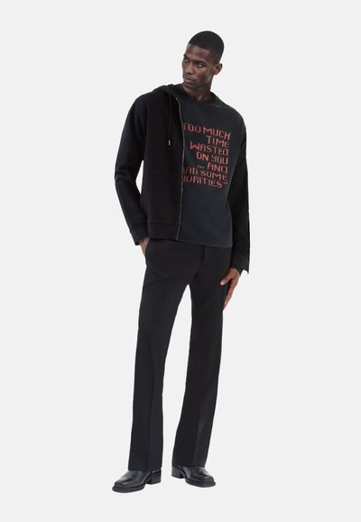 Sort lynlås hoodie over en sort sweatshirt med rødt tekst, parret med sorte bukser og sorte sko. Glat stof, afslappet pasform, minimalistisk design.