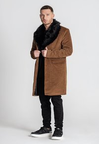Abrigo de pana marrón con cuello de piel sintética negra, dos bolsillos frontales y corte recto. Combinado con jeans negros y zapatillas.