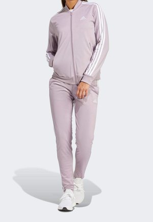 Femme portant un survêtement Adidas violet clair avec des bandes blanches, une veste zippée et des baskets blanches, marchant vers l'avant sur un fond uni.