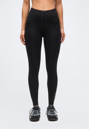 Naine, kes kannab musta kõrge vöökohaga retuuse, millel on nöörvöö ja hallid matkasaapad, seistes tavalise valge tausta ees.