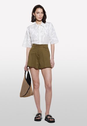 Chemise blanche à manches courtes avec des détails brodés, associée à un short vert olive. Le modèle tient un sac en osier marron et porte des sandales noires.