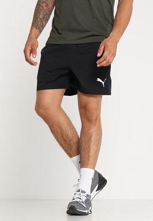 Mann trägt schwarze Puma-Shorts, graue und weiße Sneakers, weiße Socken und ein dunkelgrünes Shirt, steht mit einem Bein überkreuzt.