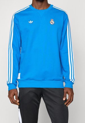 Person trägt einen blauen Adidas Real Madrid Sweatshirt mit weißen Streifen an den Ärmeln und schwarze Hose.