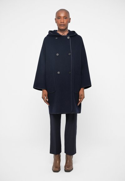 Cappotto in lana blu navy doppiopetto con cappuccio, dotato di due tasche frontali e piping a contrasto, indossato su pantaloni attillati e stivali alla caviglia marroni.