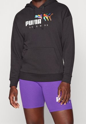 Person iført en sort Puma hættetrøje med et farverigt logo og lilla Puma shorts mod en ensfarvet baggrund.