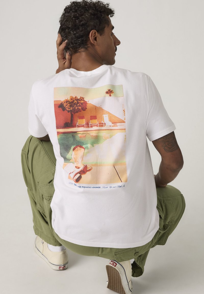 Mann mit lockigem Haar, der ein weißes T-Shirt mit Grafik und grüne Cargo-Hosen trägt, hockt und berührt mit der Hand den Kopf, dabei vom Kamera weggewandt.