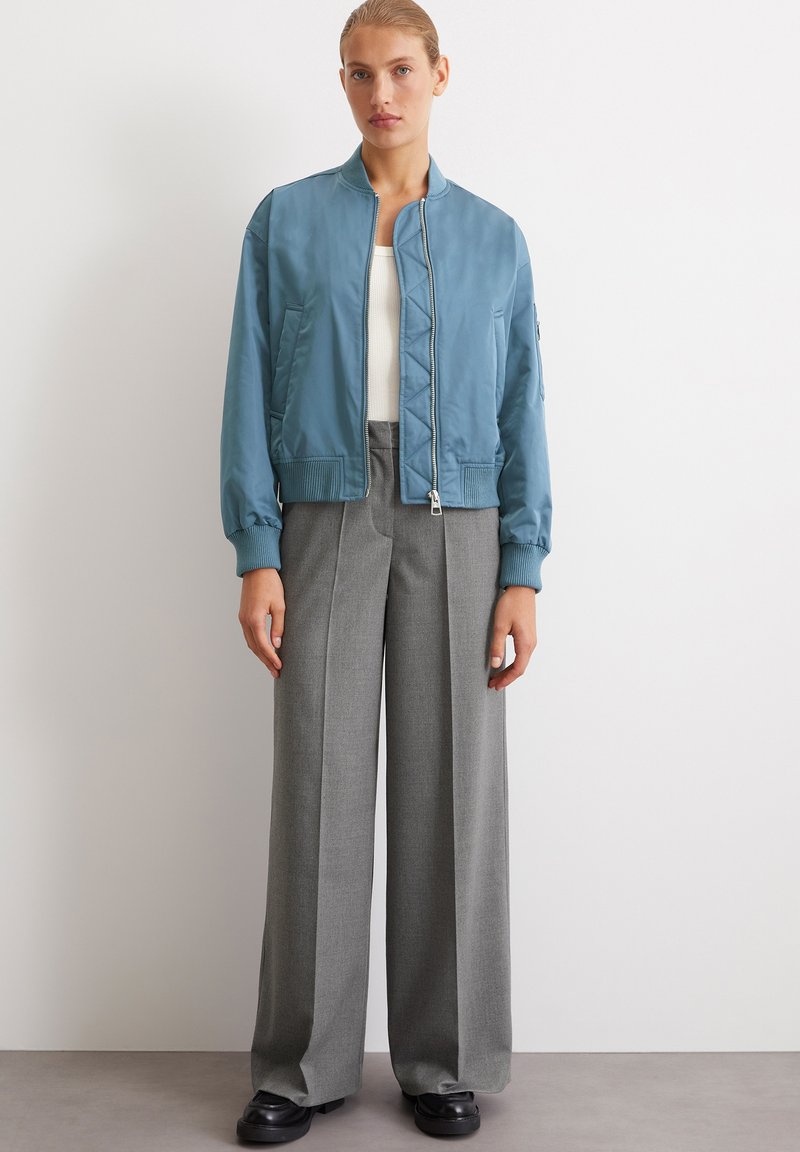 Jasnoniebieska kurtka bomberka z zapięciem na zamek, ściągaczami na rękawach oraz pikowanym wzorem, w zestawieniu z wysoko taliowanymi szarymi spodniami i czarnymi butami.