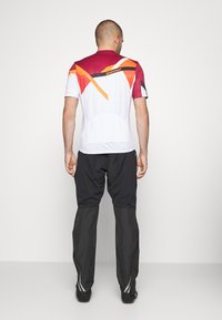 T-shirt de sport à manches courtes, principalement blanc avec des accents géométriques rouges et oranges, associé à un pantalon noir avec des bandes réfléchissantes.