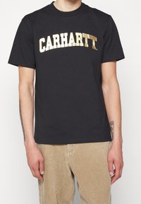Černé bavlněné tričko s výrazným metalickým zlatým nápisem "CARHARTT" na hrudi. Krátké rukávy a klasický střih s kulatým výstřihem.