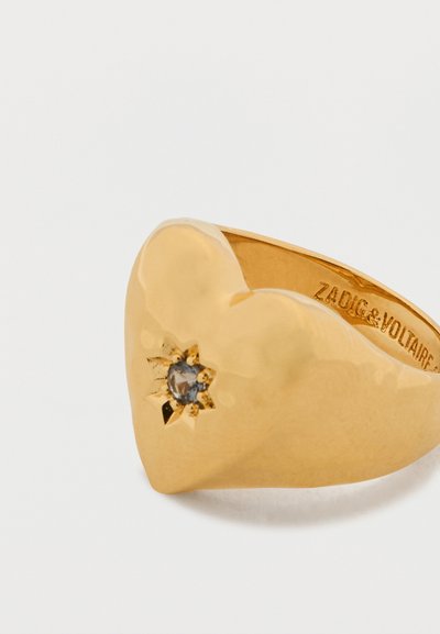 Anello a forma di cuore in oro inciso con un motivo a stelle e una piccola gemma grigia incastonata, nome del marchio inciso all'interno della fascia.