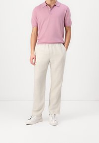 Mand iført lyserød kortærmet polo skjorte, beige løstsiddende bukser og hvide sneakers, står med den ene hånd i lommen.