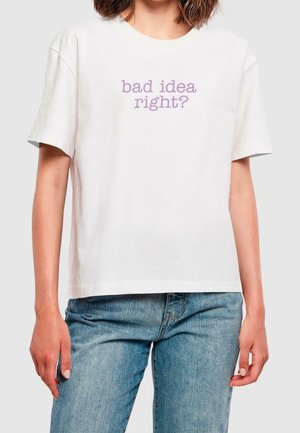 Kobieta w białej koszulce z lawendowym napisem "bad idea right?" i niebieskich dżinsach, stojąca na jednolitym jasnym tle.