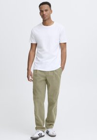 T-shirt en coton blanc associé à un chino ample de couleur vert olive clair. Le modèle porte des chaussures blanches avec des accents gris et des bandes noires.
