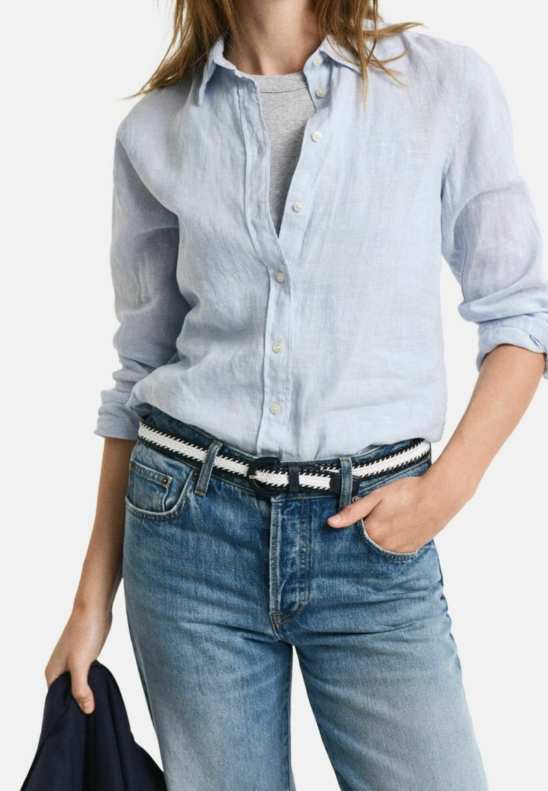 Femme portant une chemise bleu clair boutonnée sur un top gris, jean bleu avec une ceinture tressée noire et blanche, tenant une veste sombre dans une main.