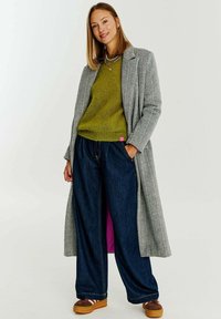 Dlouhý kabát v hřebíčkovém vzoru v šedé barvě, zelený pletený svetr, široké modré džíny, hnědé platformové boty. Růžová visačka na spodním lemu svetru.