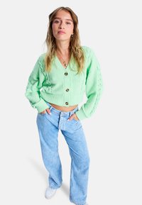 Grüne Strickjacke mit V-Ausschnitt, großen Knöpfen und Zopfmuster, kombiniert mit hellblauen weiter geschnittenen Jeans und weißen Sneakern.