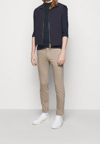 Gilet senza maniche blu, con zip, indossato sopra una maglietta scura, abbinato a pantaloni slim-fit beige e sneaker bianche. Dettaglio della zip dorata sul gilet.