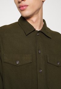 Ciemnozielona, teksturowana koszula zapinana na guziki z kołnierzykiem, dwoma przednimi kieszeniami i brązowymi guzikami, o prostym kroju i widocznych szwach.