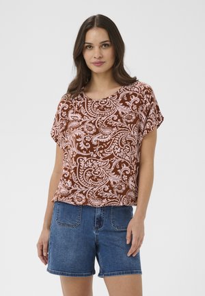 Kobieta z długimi ciemnymi włosami, ubrana w brązowy i różowy krótki top w kwiatowy wzór oraz niebieskie dżinsowe szorty, stojąca na białym tle.