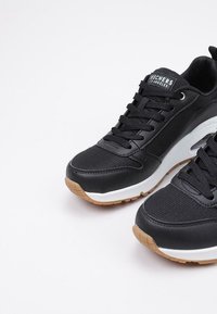 Zapatillas deportivas negras hechas de malla y materiales sintéticos, con una entresuela blanca y una suela de goma. Presentan cordones y detalles del logotipo.