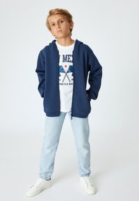 Sudadera con cremallera en color navy, con bolsillos laterales, llevada sobre una camiseta gráfica blanca que presenta banderas de cuadros azul y negro, combinada con jeans de mezclilla claros.