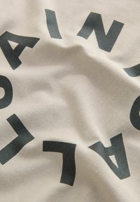 Hellbeige Stoff mit auffälligen schwarzen Buchstaben in Blockschrift, die unregelmäßige Krümmungen und Texturen aufweisen und somit ein geschichtetes Aussehen erzeugen.