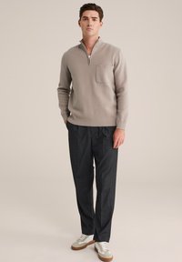 Beige Zip-Kragen-Pullover mit einer Fronttasche, kombiniert mit dunkelgrauen, maßgeschneiderten Hosen und weißen Sneakers mit einer hellbraunen Sohle.
