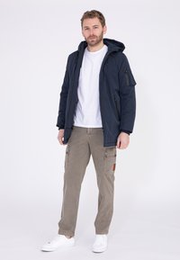 Marineblaue Kapuzenjacke mit Taschen an der Seite und an den Ärmeln, kombiniert mit hellbeigen Cargo-Hosen und weißen Sneakern. Einfaches, funktionales Design.