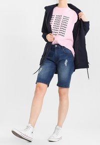 Granatowa kurtka, różowa koszulka z grafiką i powtarzającym się tekstem, dżinsowe krótki spodenki z postrzępionymi detalami oraz białe wysokie trampki.