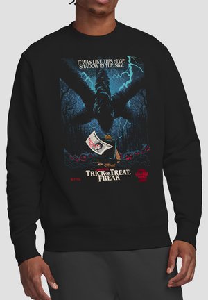 Person wearing black sweatshirt with graphic of large monster shadow, child holding sign, and text "Trick or Treat, Freak" from Stranger Things.