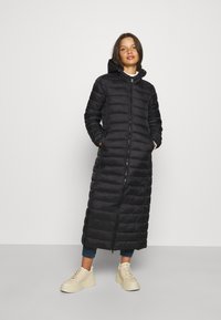 Long manteau noir matelassé avec capuche, coutures verticales, fermeture éclair avant et poches latérales, porté sur un jean décontracté et des baskets de couleur claire.