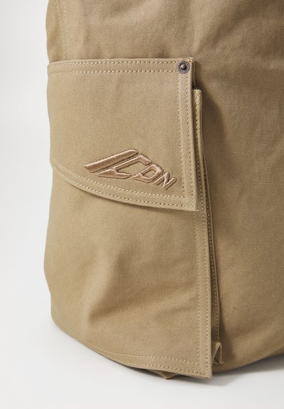 Khaki-kangasta valmistettu laukku, jossa on taittuva läppä, etutasku ja brodeerattu logo. Ompeleet ja kupariset napit antavat ilmettä.