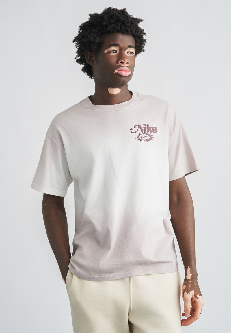 Ανοιχτό γκρι βαμβακερό μπλουζάκι με μωβ-μπουργουνδί λογότυπο Nike στο στήθος, κοντά μανίκια και χαλαρή εφαρμογή.