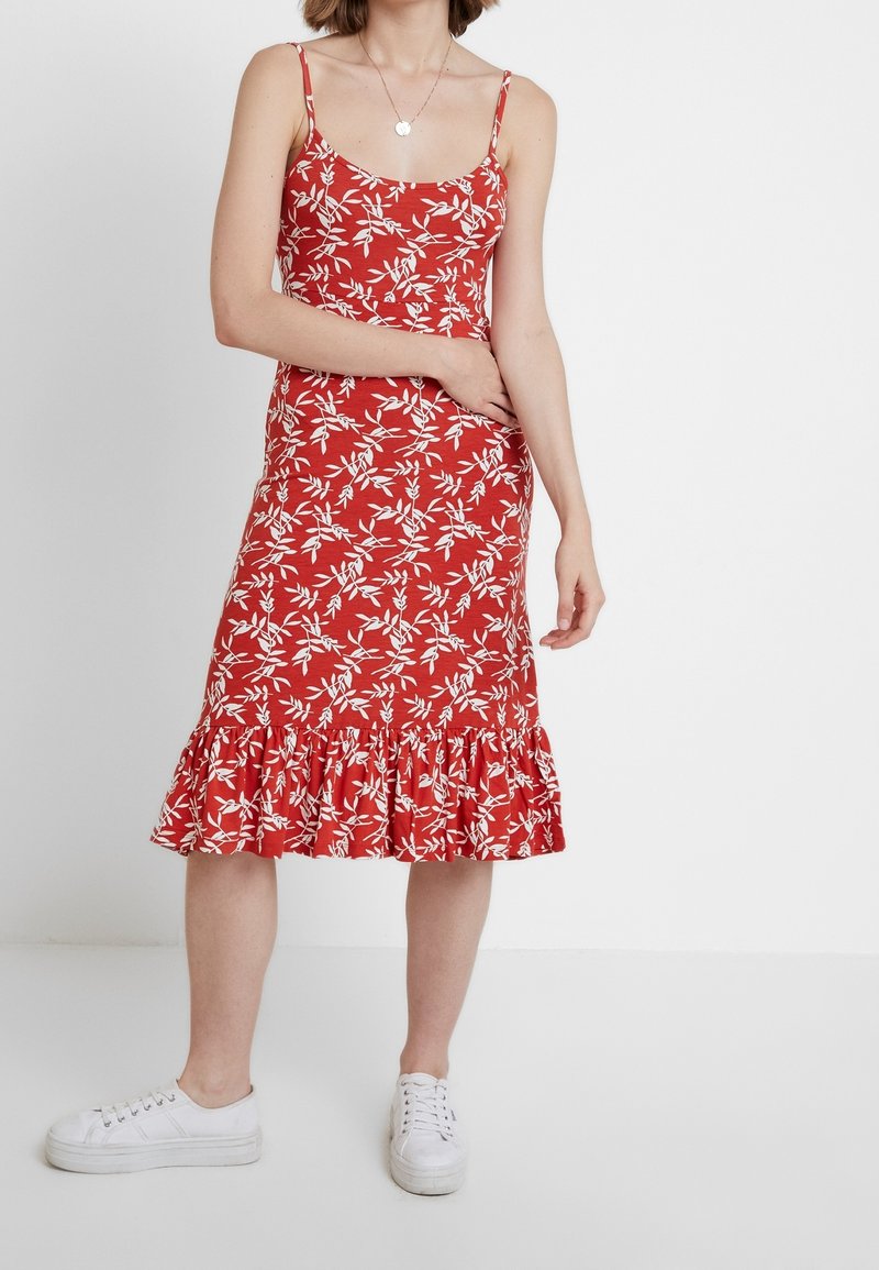 Punajuovainen kukallinen mekko, jossa on valkoisia lehtikuvioita, istuva bodice, ohuet hihnat ja röyhelöhelma. Yhdistettynä valkoisiin lenkkareihin.