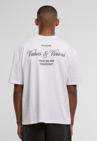 Vīrietis, kuram mugurā ir balta T-kreklu ar uzrakstiem "PEGADOR," "Values & Visions" un "Discipline and Consistency" melnā krāsā.