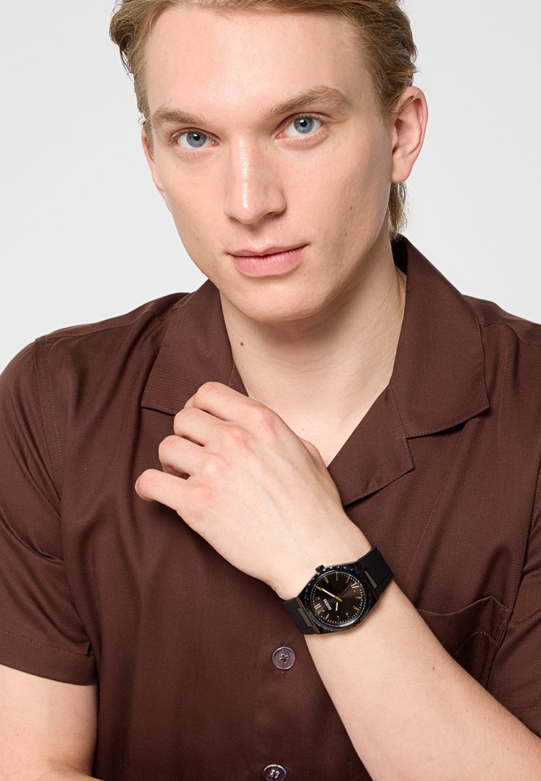 Reloj negro con esfera redonda, acentos dorados y correa de goma. Lleva puesto en la muñeca de una persona con una camisa marrón de manga corta.