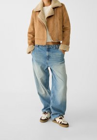 Kurtka ze skóry zamszowej w kolorze jasnobrązowym z kożuchem, o skróconym kroju, zestawiona z jasnoniebieskimi spodniami o szerokich nogawkach i wzorzystymi trampkami.