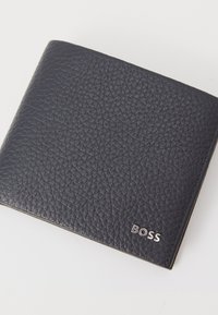 Portafoglio in pelle nera con superficie texturizzata, caratterizzato da un logo "BOSS" in argento nell'angolo inferiore destro. Forma compatta e quadrata.