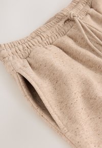 Gros plan sur un pantalon de survêtement beige en maille avec taille élastique, cordon, poche latérale et texture de tissu mouchetée.