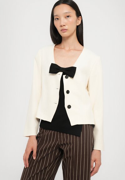 Cream blazer with black bow accent, five black buttons, and a cropped design. Features long sleeves and a rounded neckline.